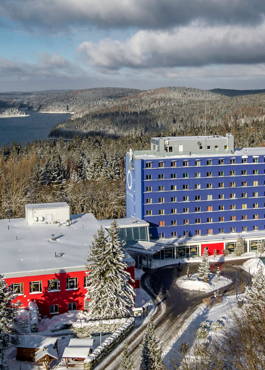 Luftaufnahme des Familienhotels am Bühl in Eibenstock mit mehrstöckigem blauen Hauptgebäude, rotem Nebengebäude, Garten mit Sitzgruppen und Blick auf bewaldete Hügel und einen See im Erzgebirge