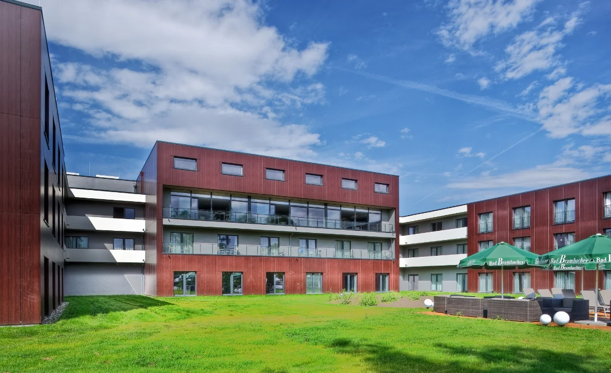 Moderne Gebäudefassade des Sante´Royale in Bad Langensalza mit klaren Linien und großen Fensterflächen. Die Fassade besteht aus rötlich-braunen Paneelen und weißen Elementen. Im Vordergrund erstreckt sich eine gepflegte Rasenfläche. Rechts stehen mehrere grüne Sonnenschirme mit weißer Aufschrift über einer Terrasse mit Sitzgelegenheiten. Der Himmel ist blau mit vereinzelten Wolken.
