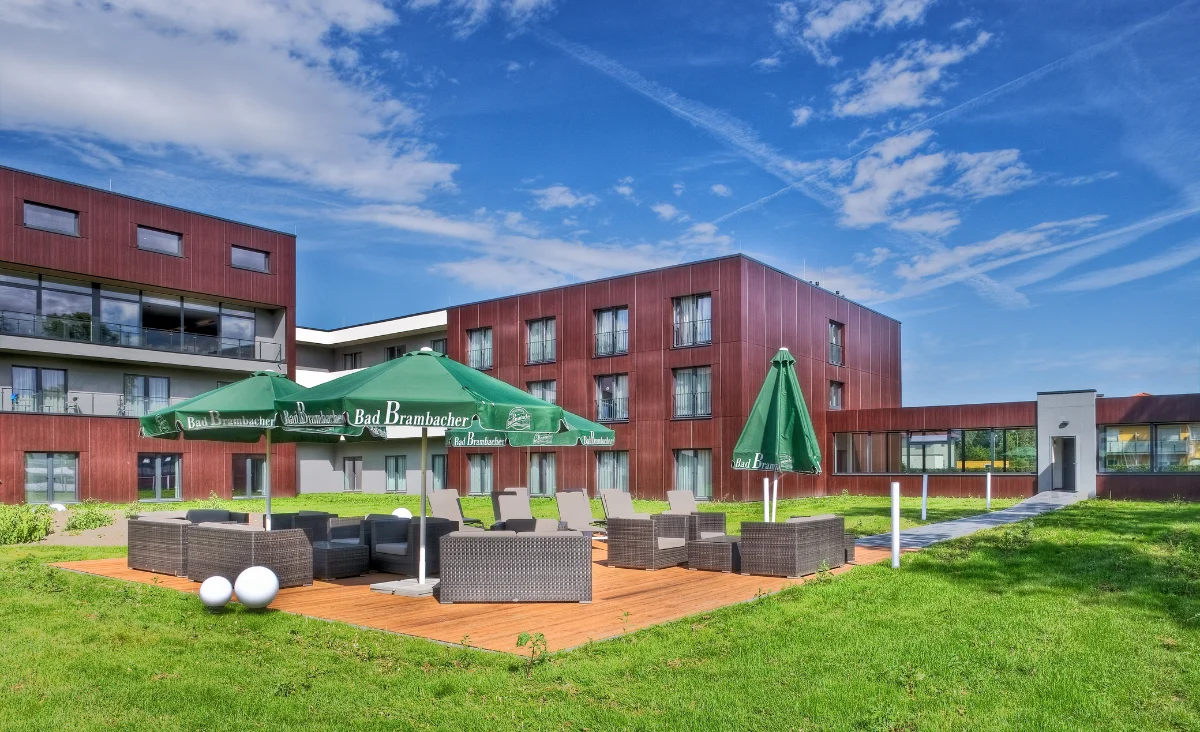 Moderne Gebäudefassade des Sante´Royale in Bad Langensalza mit klaren Linien und großen Fensterflächen. Die Fassade besteht aus rötlich-braunen Paneelen und weißen Elementen. Im Vordergrund erstreckt sich eine gepflegte Rasenfläche. Rechts stehen mehrere grüne Sonnenschirme mit weißer Aufschrift über einer Terrasse mit Sitzgelegenheiten. Der Himmel ist blau mit vereinzelten Wolken.