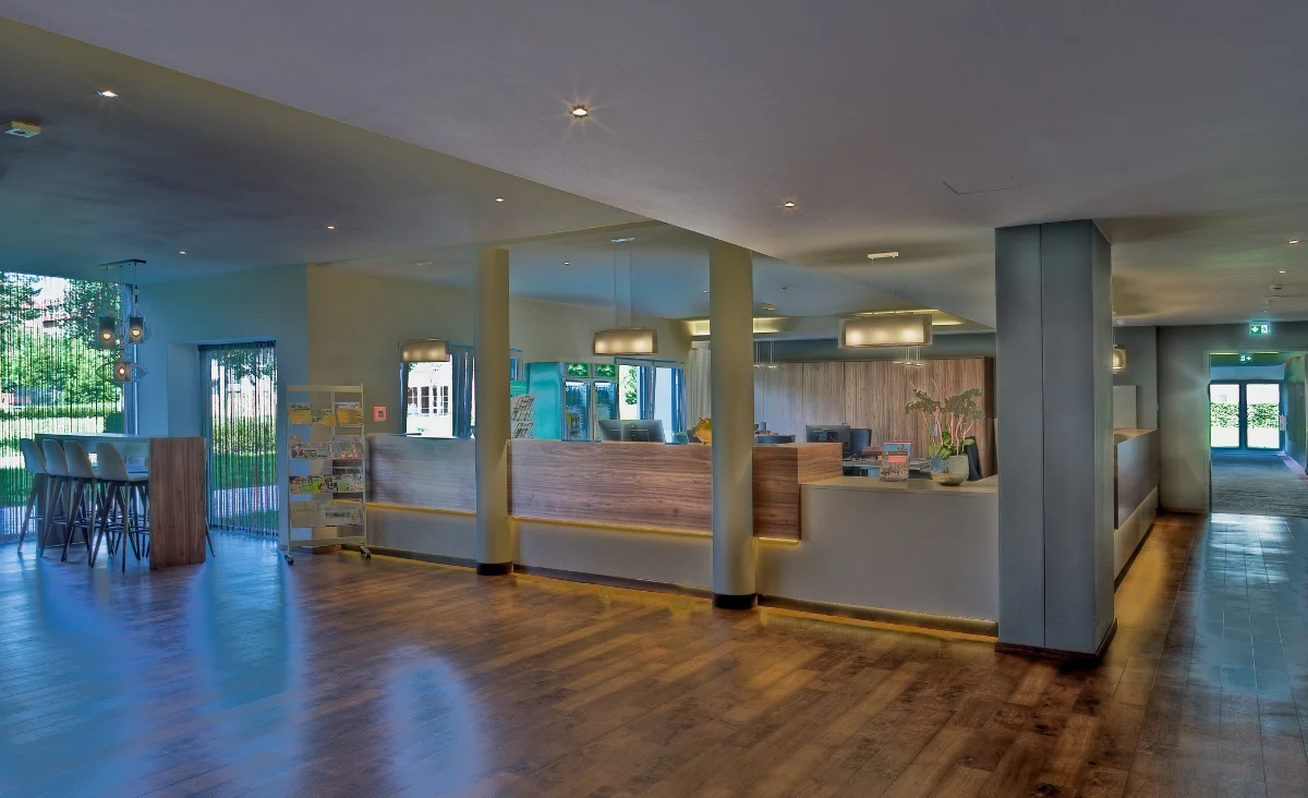 Großzügiger, modern gestalteter Empfangsbereich des Sante´Royale in Bad Langensalza mit hellem Holzfußboden und dezent beleuchteter Rezeption. Die Theke ist in mehreren Segmenten angeordnet, mit hellen Holzverkleidungen und weißen Oberflächen. Im Hintergrund befinden sich Sitzgelegenheiten und ein Zeitschriftenständer. Große Fensterfronten lassen Tageslicht herein, Pflanzen und dekorative Leuchten sorgen für eine einladende Atmosphäre. Rechts führt ein breiter Flur zu einer Glastür.