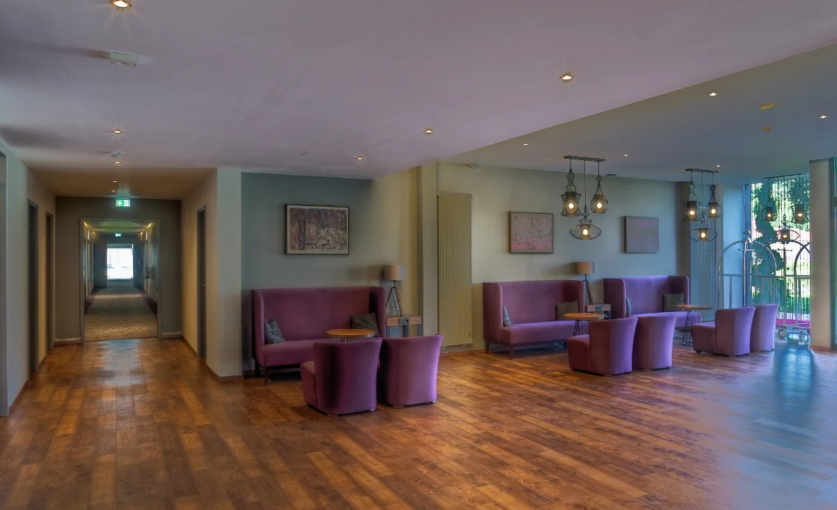 Im Vordergrund erstreckt sich ein großzügiger Aufenthaltsbereich mit mehreren lilafarbenen Sofas und Sesseln, die um niedrige Tische gruppiert sind. Die Sitzgruppen stehen auf einem Boden in Holzoptik. An den Wänden hängen gerahmte Bilder. Mehrere moderne Deckenleuchten mit Glaslampenschirmen sorgen für Beleuchtung. Rechts fällt Tageslicht durch große Fenster und eine Glastür, die den Blick auf eine grüne Außenanlage freigeben. Links führt ein langer Flur mit Türen zu weiteren Räumen. Die Szene vermittelt den Eindruck eines einladenden, gepflegten Hotel- oder Klinikfoyers im Sante´Royale Bad Langensalza.