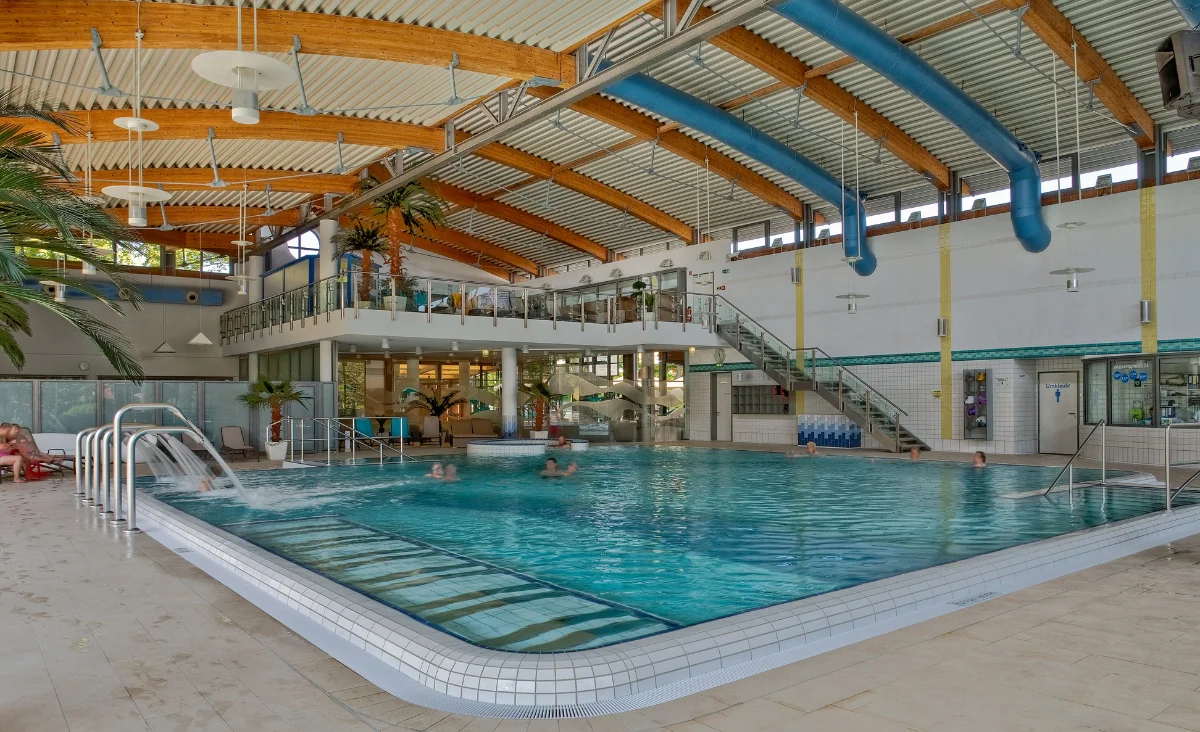 Im Vordergrund erstreckt sich ein großes, rechteckiges Schwimmbecken mit hellblauen Fliesen und einem breiten, flachen Einstiegsbereich. Mehrere Personen befinden sich im Wasser. Im Hintergrund sind eine erhöhte Galerie mit Glasgeländer, eine breite Treppe und eine Rutsche zu sehen. Die Decke besteht aus gebogenen Holzbalken und ist mit großen, blauen Lüftungsrohren versehen. Palmen und große Fenster sorgen für eine helle, freundliche Atmosphäre. Das Bild zeigt einen Innenbereich des Sante´Royale in Bad Langensalza.