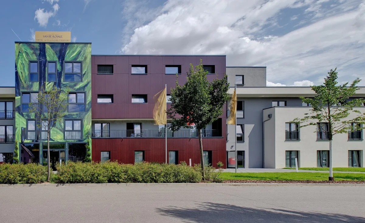 Moderne Gebäudefassade des Sante´Royale in Bad Langensalza mit klaren Linien und großen Fensterflächen. Die Fassade besteht aus rötlich-braunen Paneelen und weißen Elementen. Im Vordergrund erstreckt sich eine gepflegte Rasenfläche. Rechts stehen mehrere grüne Sonnenschirme mit weißer Aufschrift über einer Terrasse mit Sitzgelegenheiten. Der Himmel ist blau mit vereinzelten Wolken.