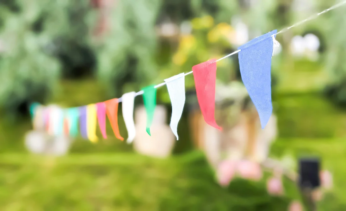 Bunte Wimpelgirlande aus Stoff hängt quer im Vordergrund und verläuft von links nach rechts. Im Hintergrund ist unscharf eine grüne Gartenlandschaft mit Bäumen und Sträuchern zu erkennen. Die Szene vermittelt eine sommerliche, festliche Atmosphäre, passend zum Tag des Gartens Neukölln.