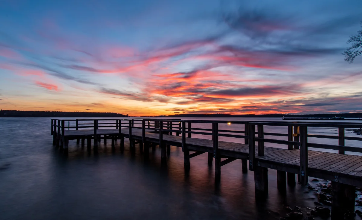 Holzsteg mit Geländer über ruhigem Wasser bei Sonnenuntergang mit farbigem Himmel und Wolken