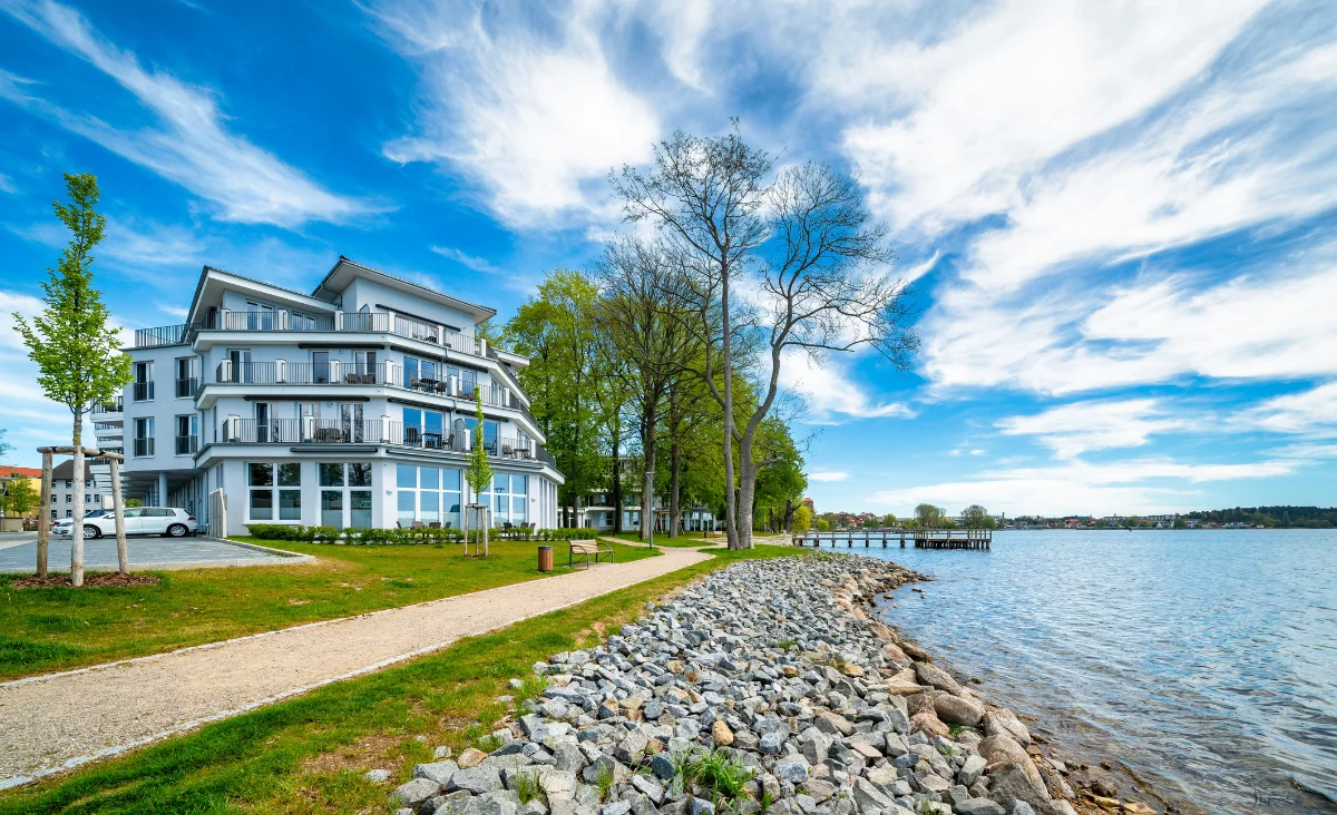 Modernes Aparthotel mit weißer Fassade und großen Fenstern am Ufer eines Sees mit steinigem Ufer und angrenzendem Fußweg