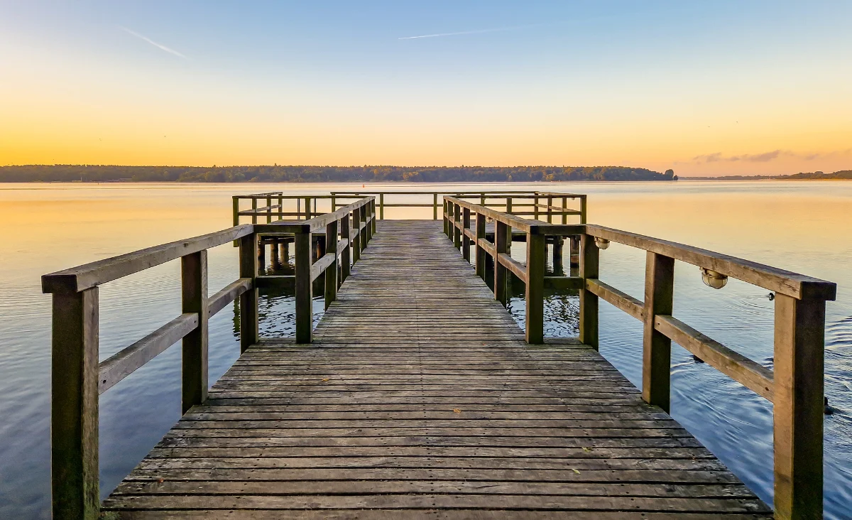 Holzsteg mit Geländer führt über ruhiges Wasser bei Sonnenuntergang