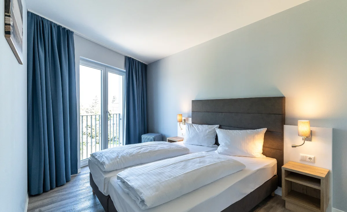 Hotelzimmer mit zwei Einzelbetten, grauem gepolstertem Kopfteil, Nachttischen mit Wandlampen und bodentiefen Fenstern mit blauen Vorhängen