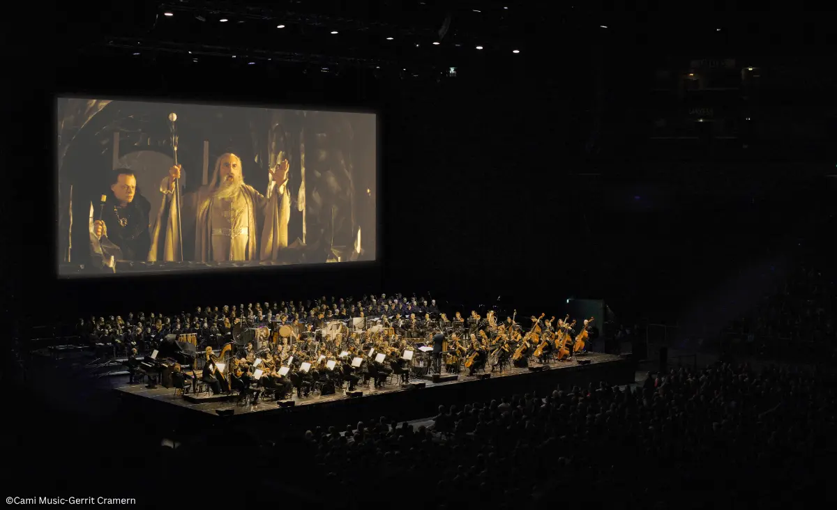 Orchester und Chor spielen vor einer Leinwand, auf der eine Szene aus 'Der Herr der Ringe: Die zwei Türme' mit zwei Figuren in mittelalterlicher Kleidung zu sehen ist