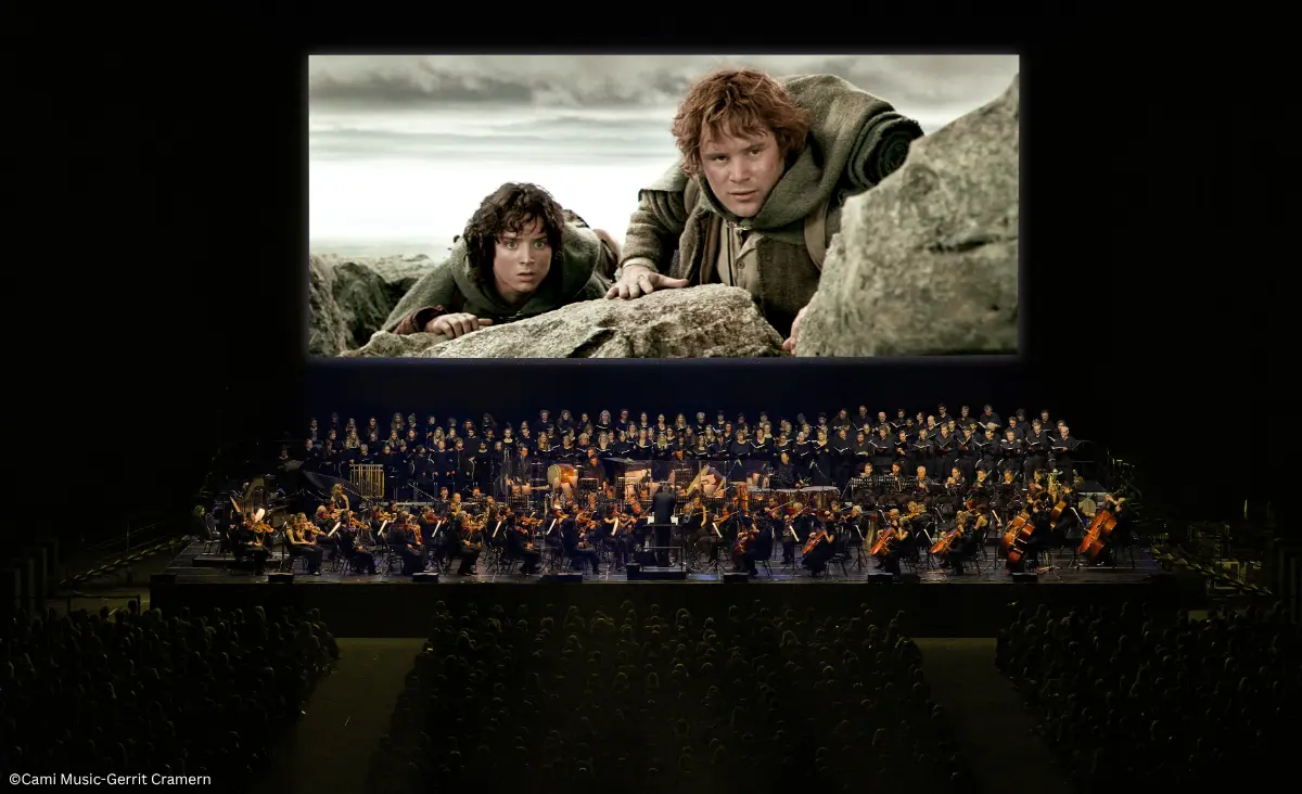 Konzertsaal mit Orchester und Chor vor großer Leinwand, die eine Szene aus "Der Herr der Ringe: Die zwei Türme" mit Frodo und Samweis.