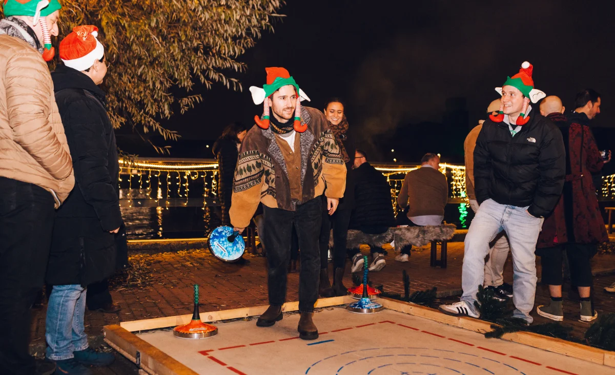 Gäste beim winterlichen Eisstockschießen im Freien an der beleuchteten Spreeterrasse während des All Inclusive Weihnachtsmarkt Schöneweide