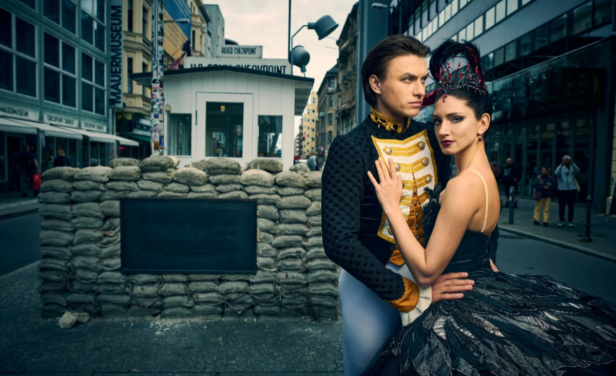 Ein Paar in klassischer Ballettkleidung steht vor einem mit Sandsäcken gesicherten Straßenschild mit der Aufschrift 'Check Point Charlie' in einer urbanen Straßenszene.
